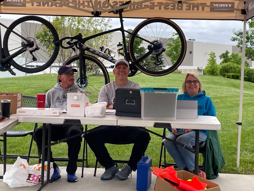 3 ABD members, seen sitting under a tent,  fill some of our Volunteer Opportunities at a time trial race check-in table.