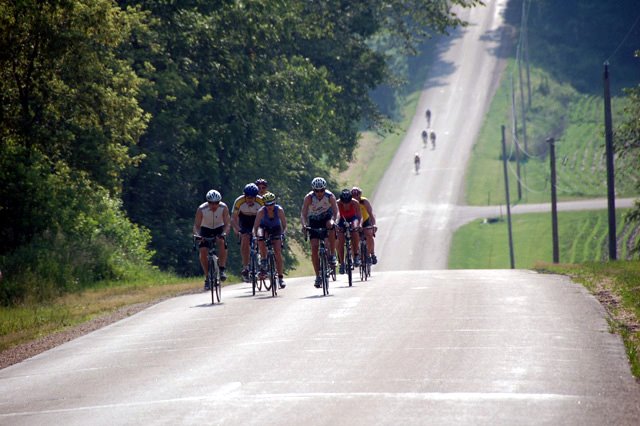 A pack of riders climbs a hilly section of the L'Alpe Bl'Huez Hill Climb.  In the distance, a smaller set of riders looks to narrow the gap.